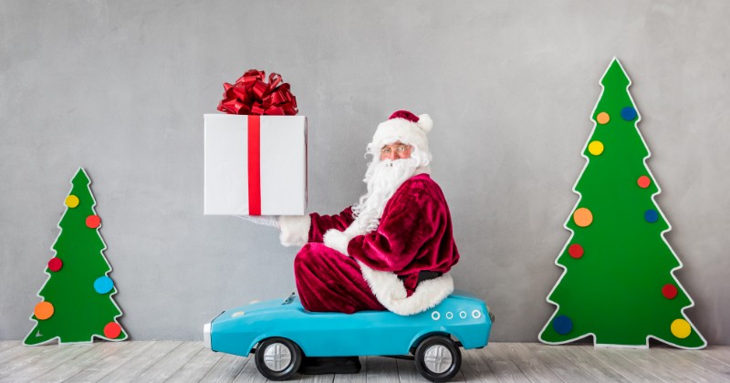 Père Noël tenant un cadeau, assis dans une petite voiture devant deux sapins de Noël